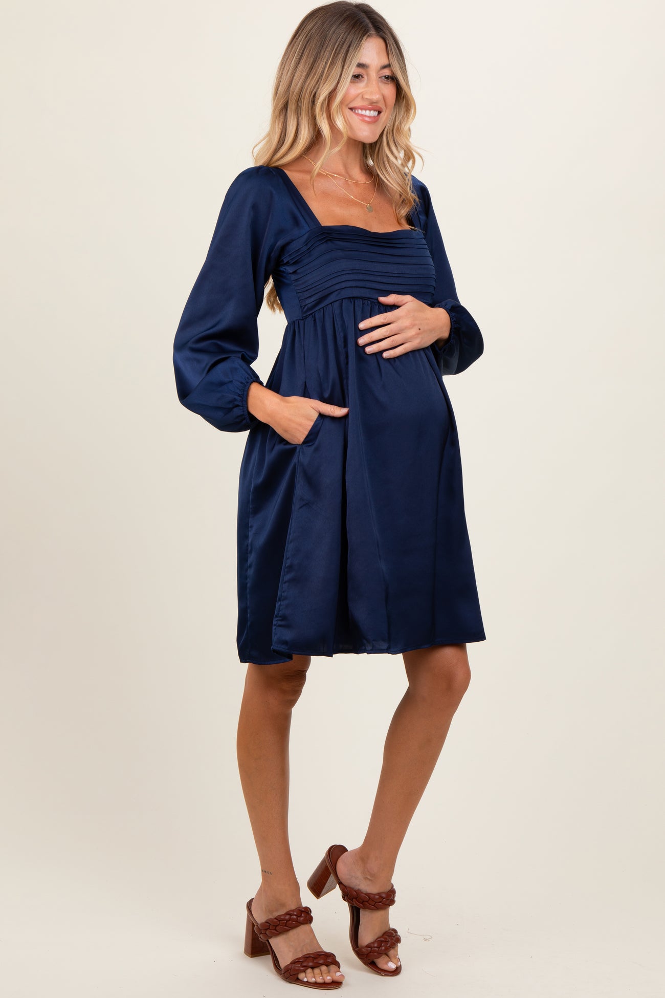 woman wearing a navy blue satin maternity dress with a square neckline and long sleeves, holding her baby bump