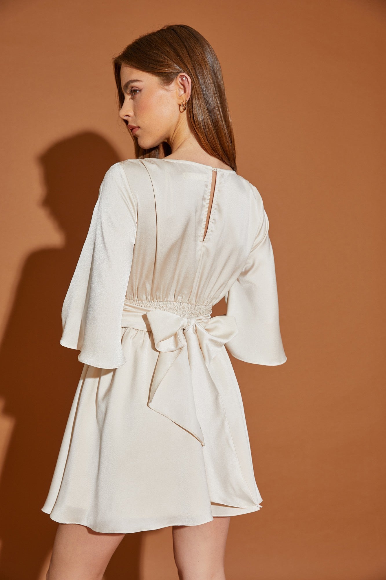 woman wearing a cream colored dress with bell sleeves and a tied waist, shown from the back against a tan background