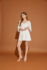 woman wearing a cream short sleeve wrap dress with a v neckline and flared sleeves, standing against a brown backdrop