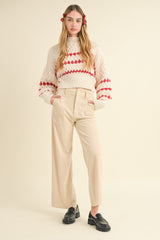 woman wearing a cream cable knit sweater with red accents and high-waisted beige wide-leg pants