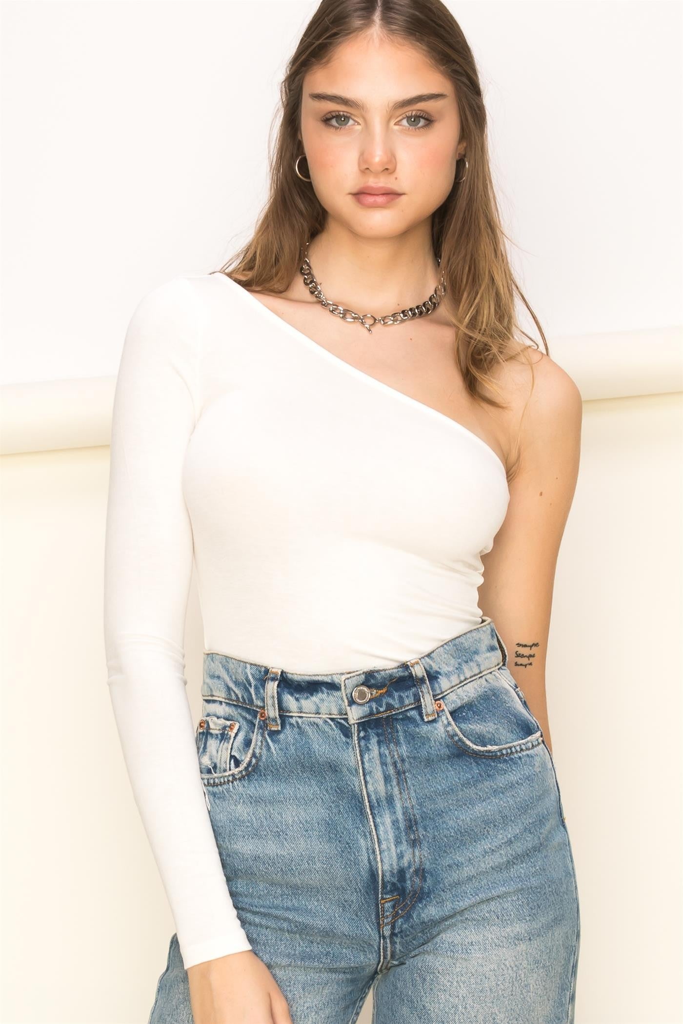 woman wearing a white one-shoulder fitted top with a long sleeve, paired with high-waisted denim jeans
