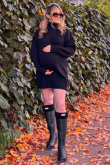woman wearing a black maternity sweater dress and black knee-high rain boots, smiling and holding her baby bump
