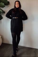 woman wearing a black oversized sweater dress cinched at the waist with a belt, paired with black tights and slippers