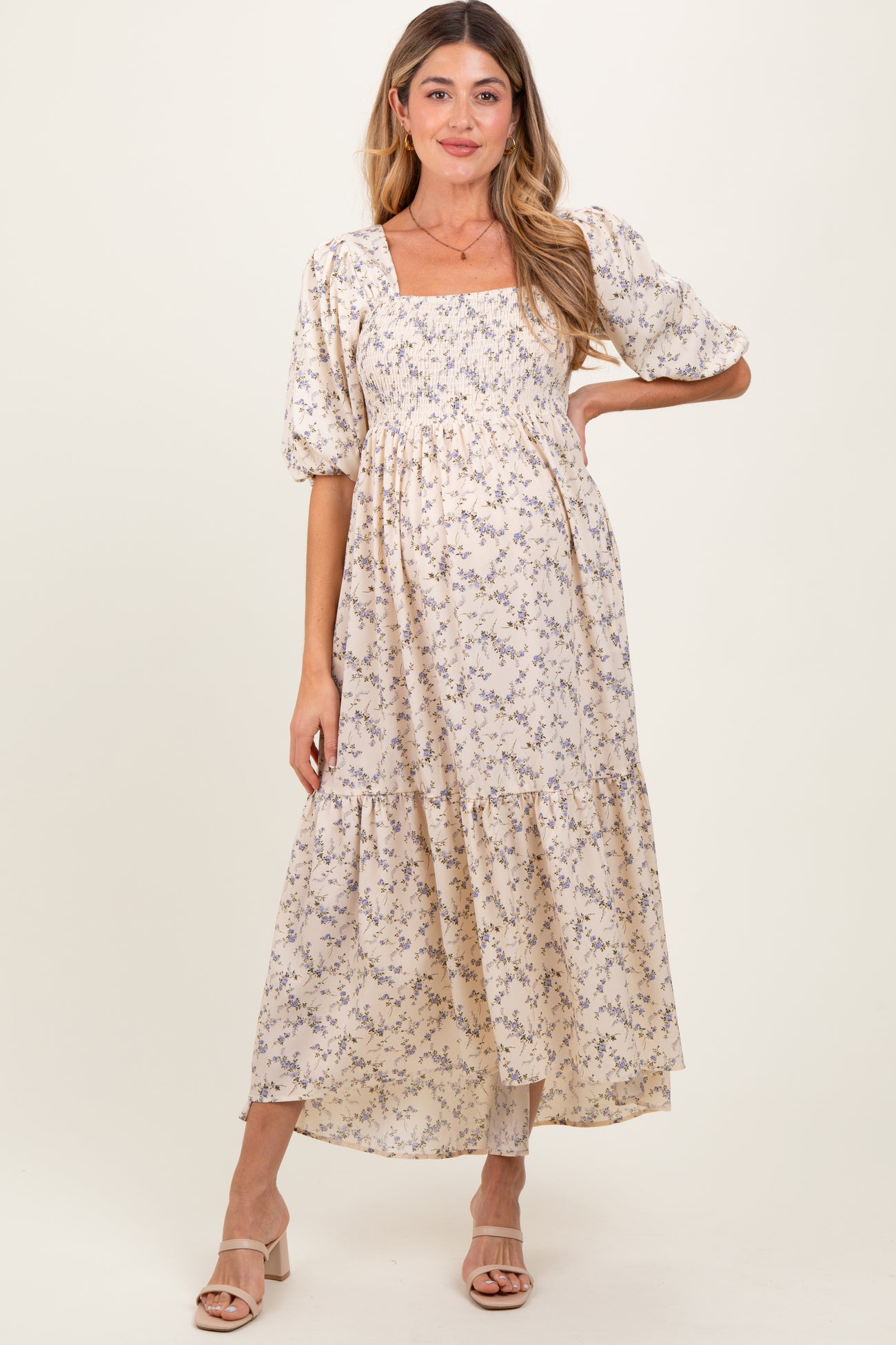 woman wearing a light beige floral maternity dress with puff sleeves and tiered skirt, showcasing her baby bump