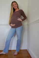 woman wearing a brown ribbed long sleeve maternity top and blue jeans, holding her baby bump