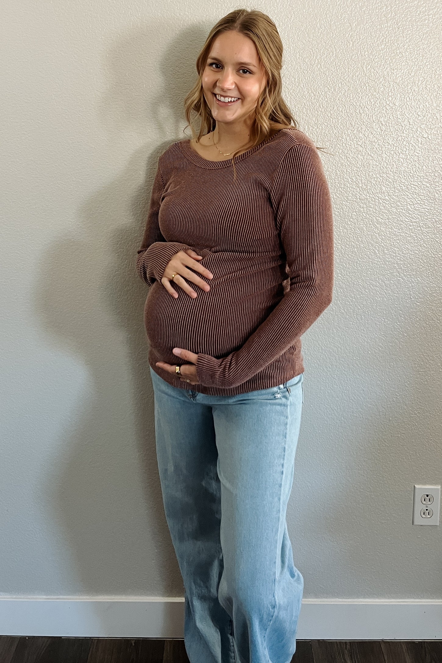 woman wearing a brown ribbed long sleeve maternity top and blue flared jeans, holding her baby bump