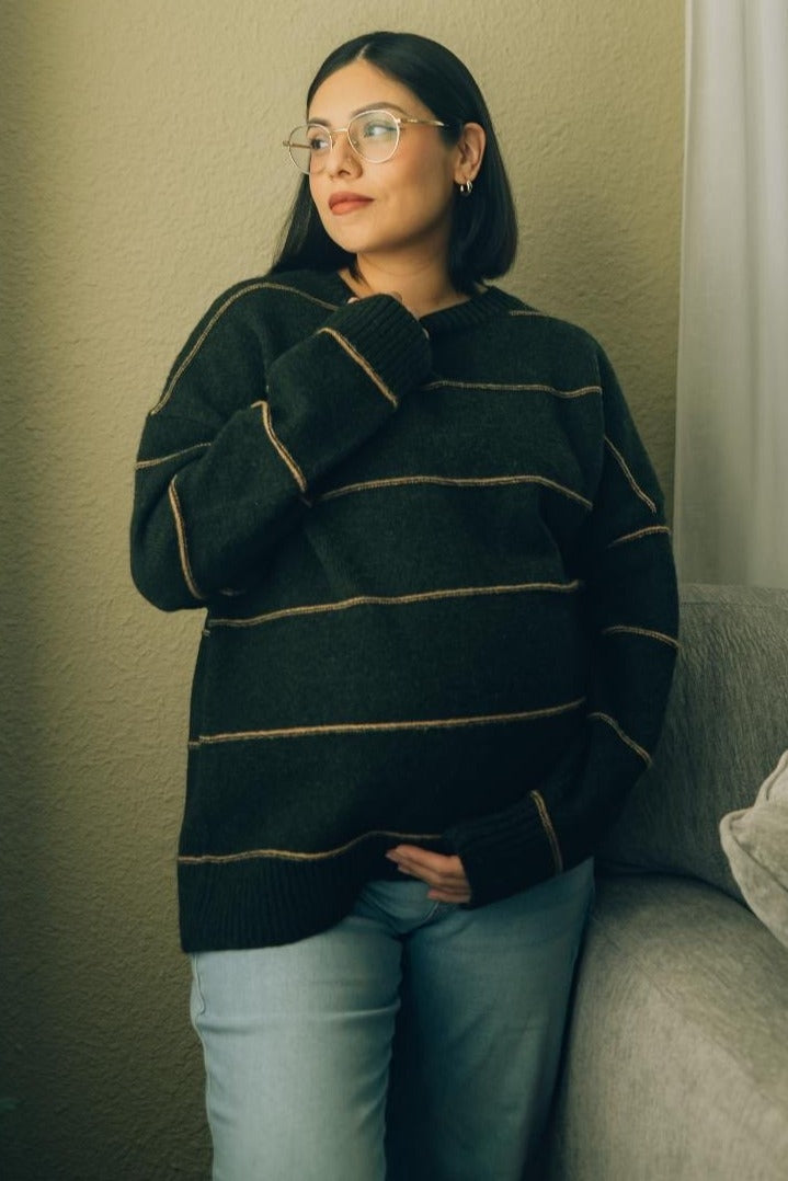 woman wearing a dark green striped maternity sweater and light blue jeans, gently holding her baby bump