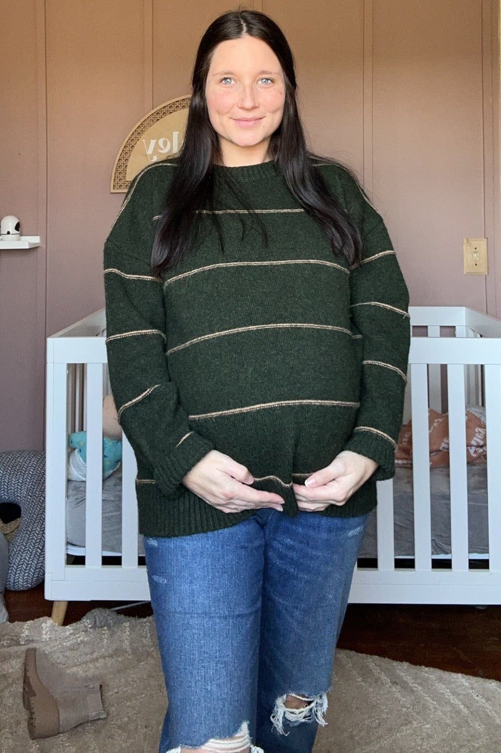 woman wearing a dark green striped maternity sweater and showing her baby bump while standing in front of a crib