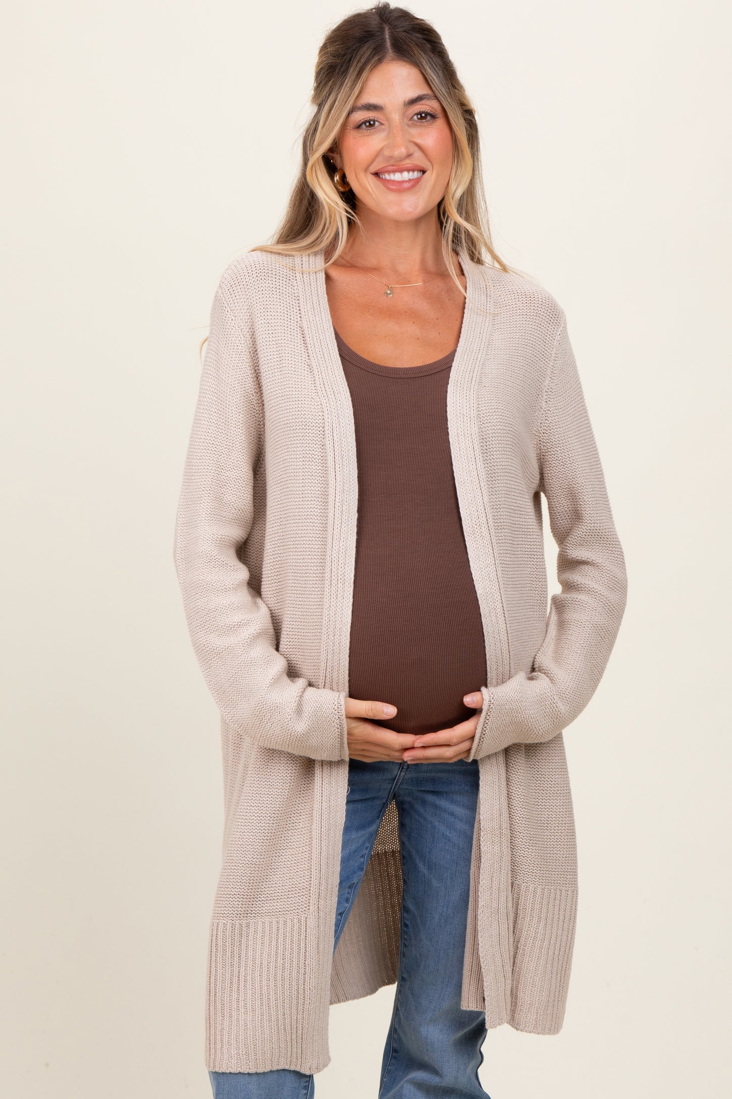 woman wearing a beige long cardigan over a brown tank top, showing her baby bump in maternity wear