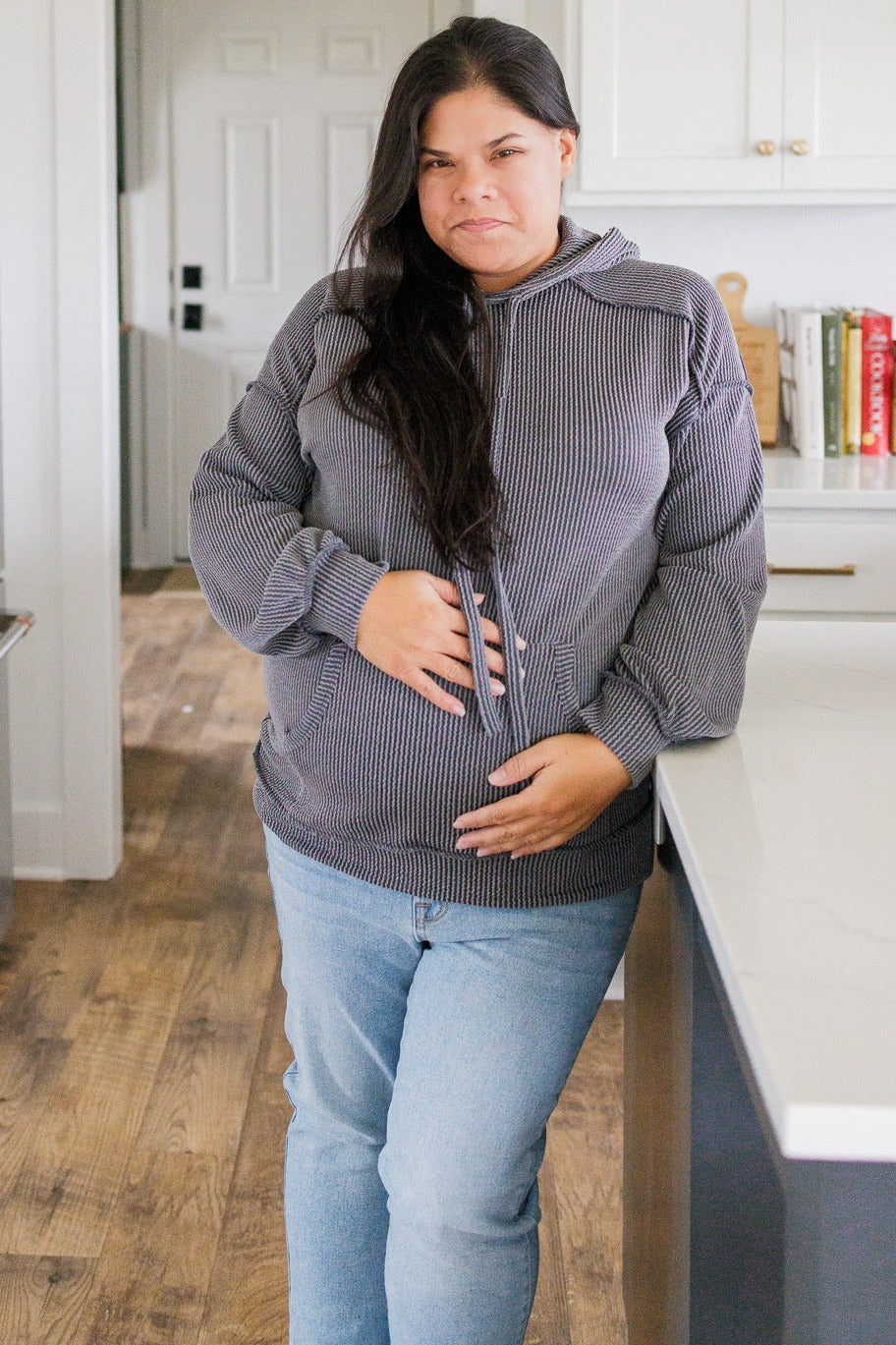 woman wearing a gray striped maternity hoodie and holding her baby bump, paired with light blue jeans