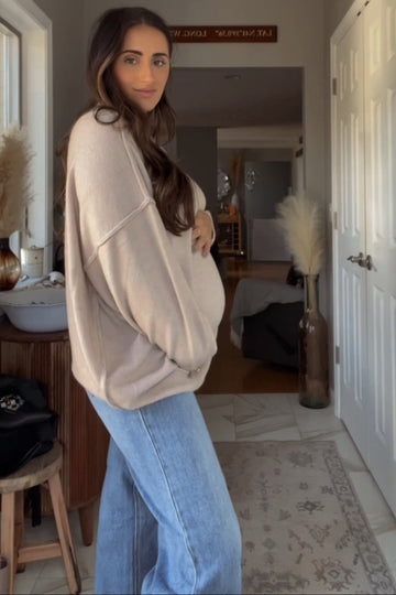 woman wearing a beige oversized maternity sweater and blue jeans, holding her baby bump in a cozy home setting