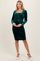 woman wearing a dark green velvet maternity dress with long sleeves and gathered detailing, showing her baby bump