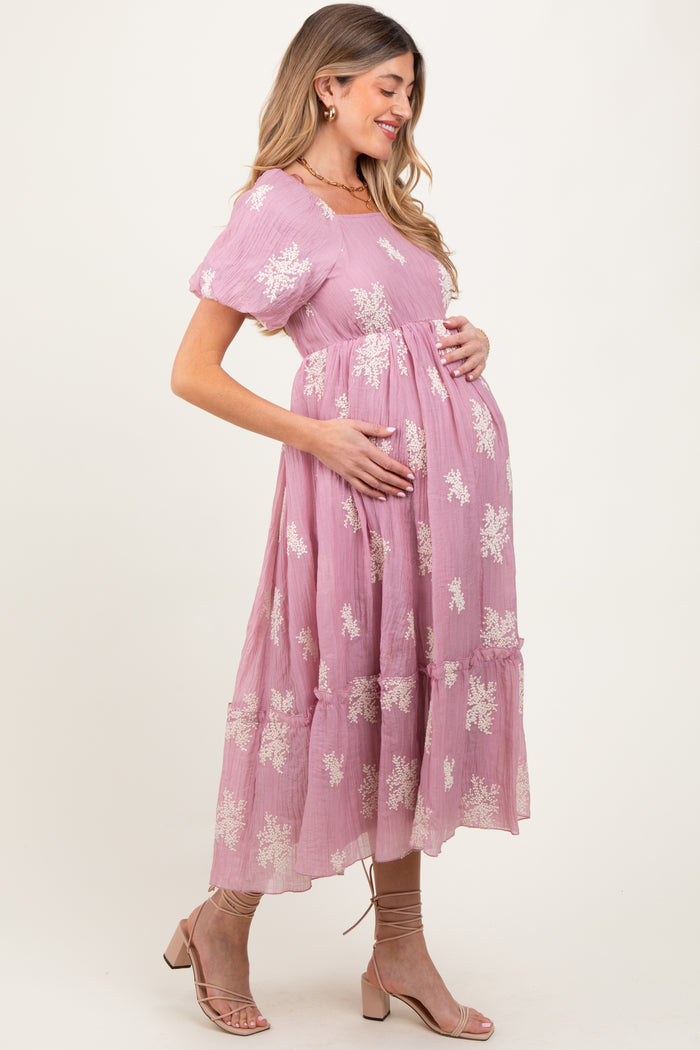 woman wearing a pink maternity dress with floral embroidery and a tiered skirt, gently cradling her baby bump