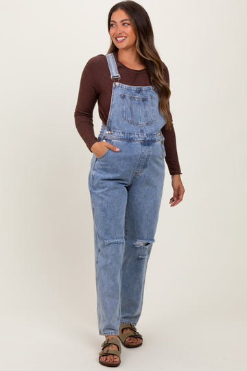woman wearing a brown long sleeve top and blue distressed maternity overalls, smiling with hands in pockets