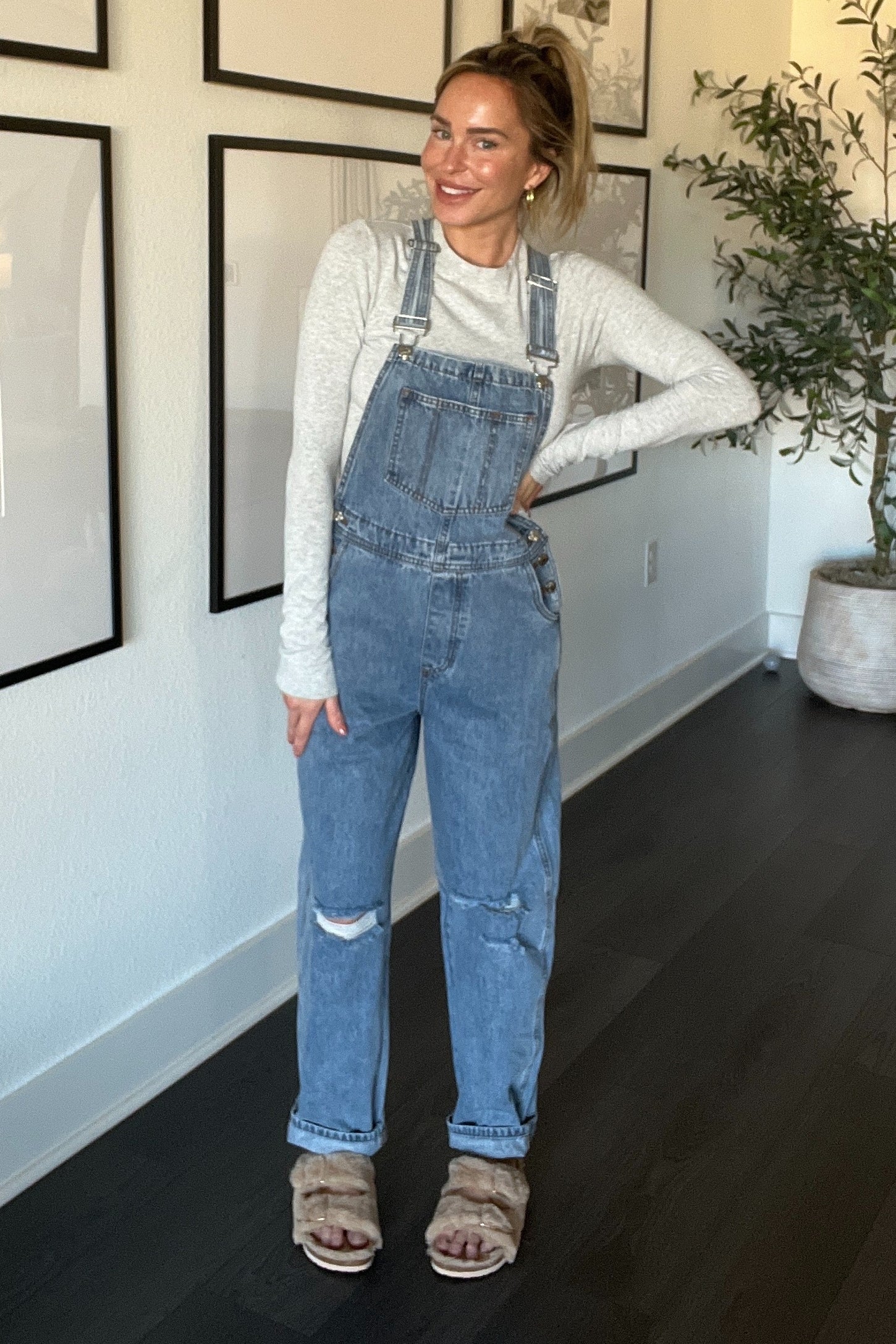 woman wearing light blue denim overalls with ripped details and a long sleeve gray top, styled with fuzzy slides