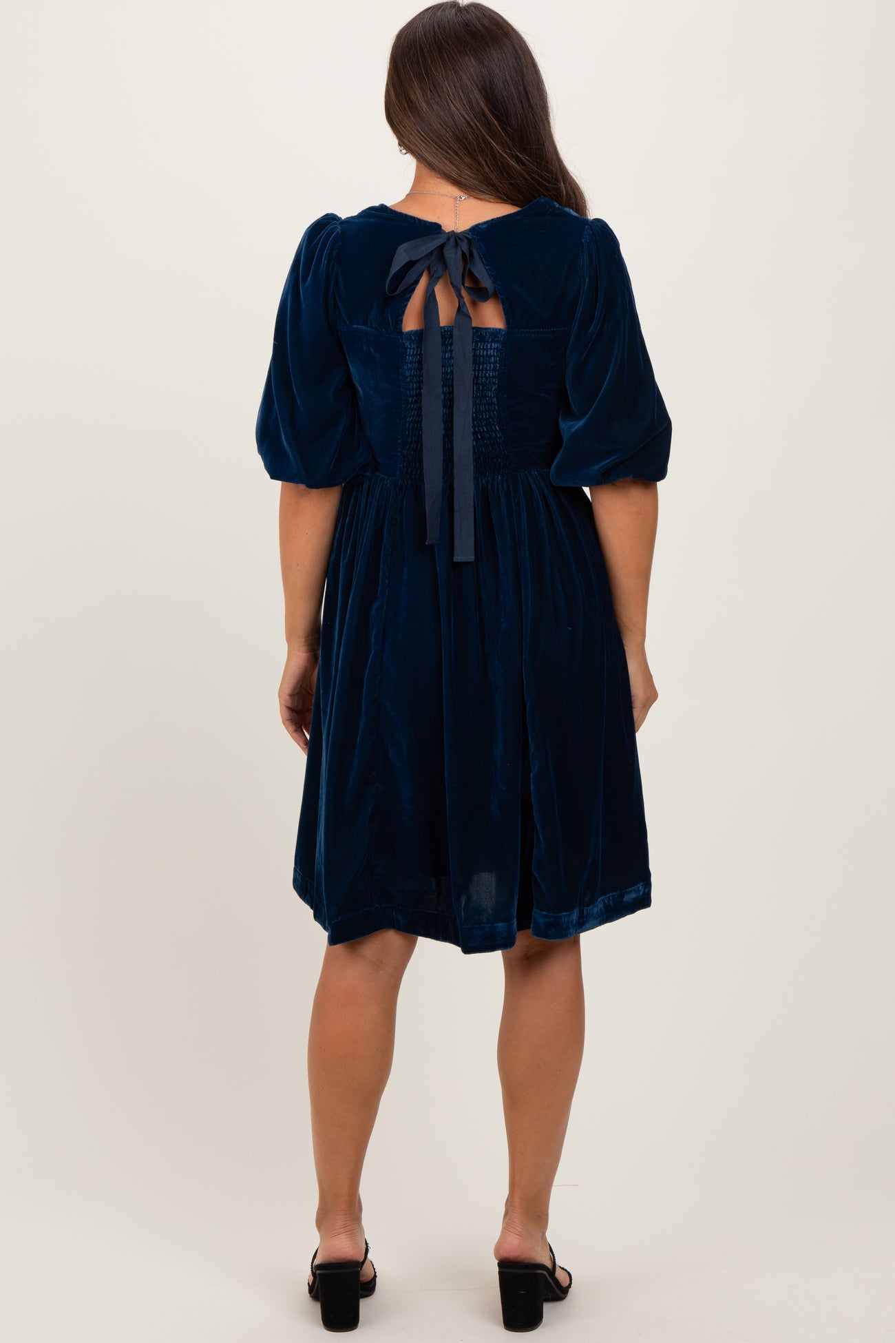woman wearing a navy blue velvet maternity dress with a smocked back and bow detail, showing off her baby bump