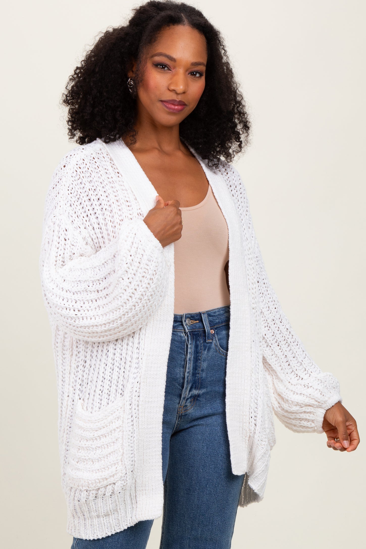 woman wearing a white oversized cardigan with textured knitting and pockets, paired with a beige tank top and denim jeans