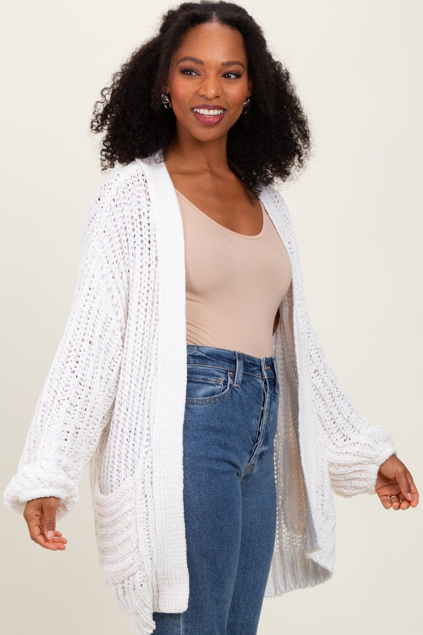 woman wearing a light beige fitted top and an oversized white knitted cardigan over high-waisted blue jeans