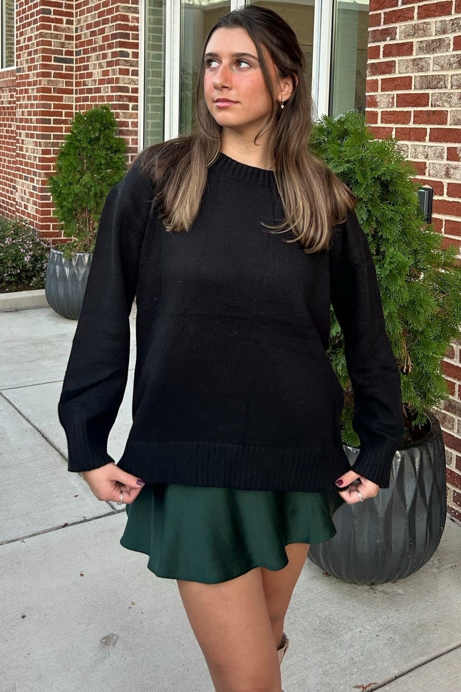 woman wearing a black oversized sweater paired with a green skirt, posing outdoors in front of planters