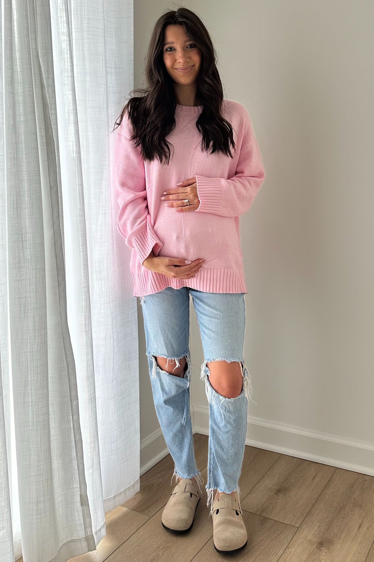 woman wearing a light pink maternity sweater and distressed jeans, holding her baby bump while standing indoors
