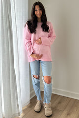 woman wearing a light pink maternity sweater and distressed jeans, holding her baby bump while standing indoors