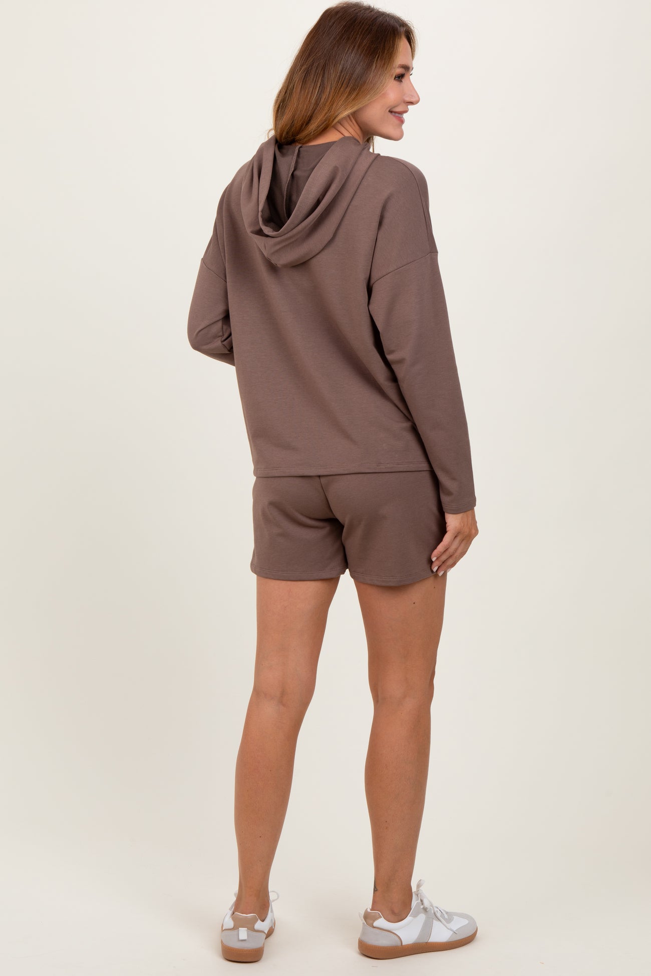 woman wearing a brown hoodie with shorts, showing the back view and paired with white sneakers