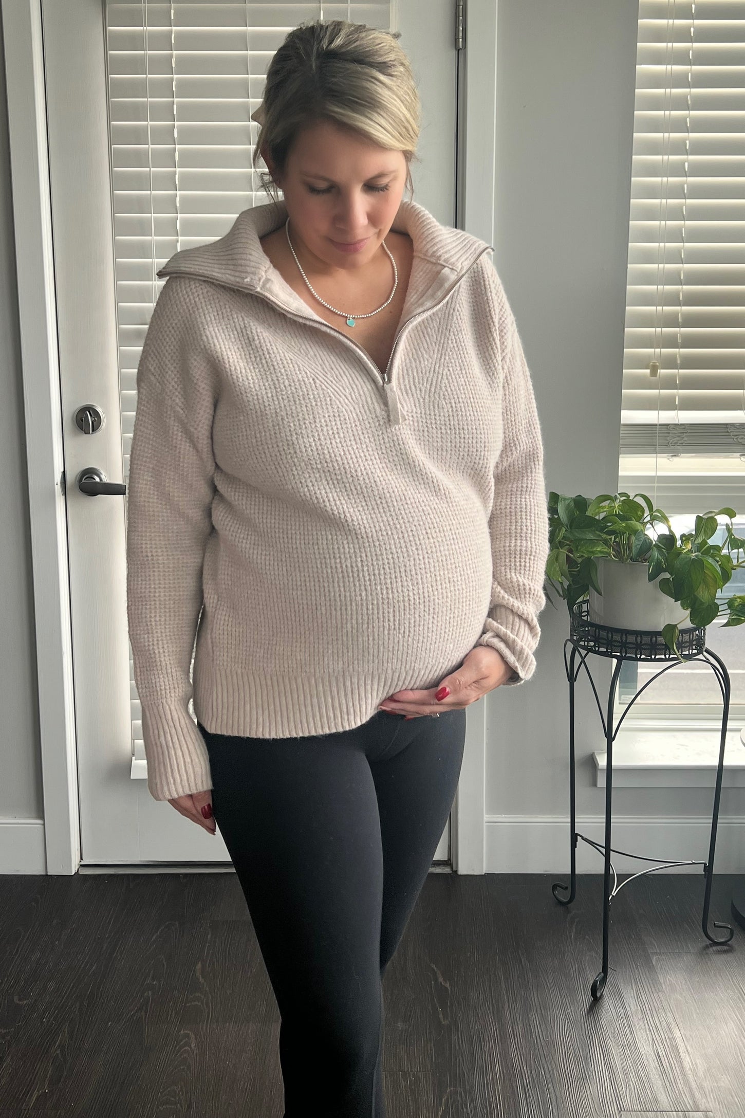woman wearing a light beige maternity pullover sweater, holding her baby bump, paired with black leggings