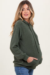 woman wearing a dark green ribbed maternity hoodie with a kangaroo pocket, gently cradling her baby bump