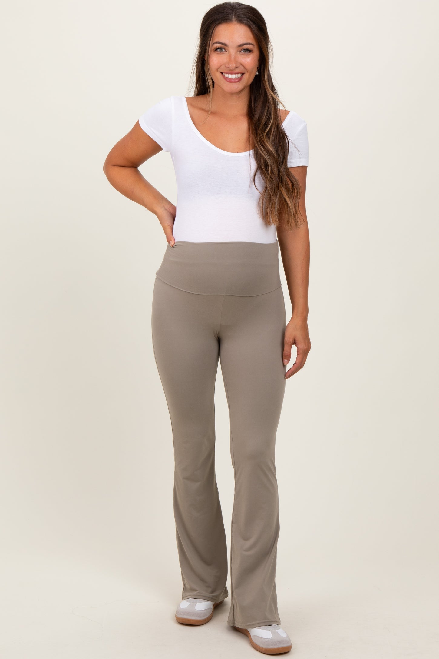 woman wearing a white scoop neck maternity top and beige maternity flared leggings, smiling and standing with one hand on her hip
