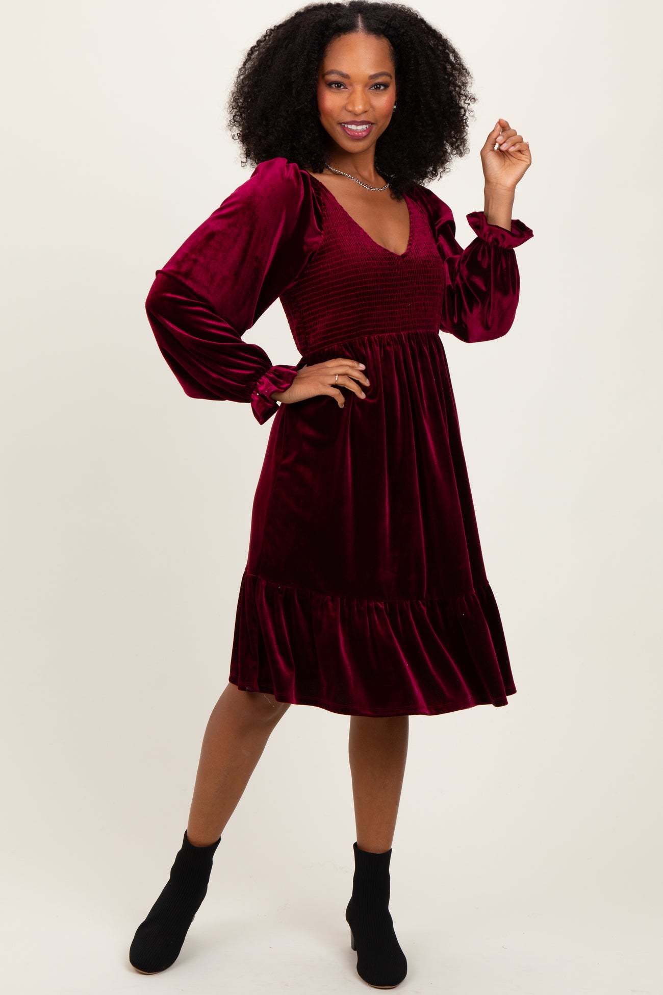 woman wearing a burgundy velvet smocked dress with long puff sleeves and a flared hem, posing with one hand on her hip