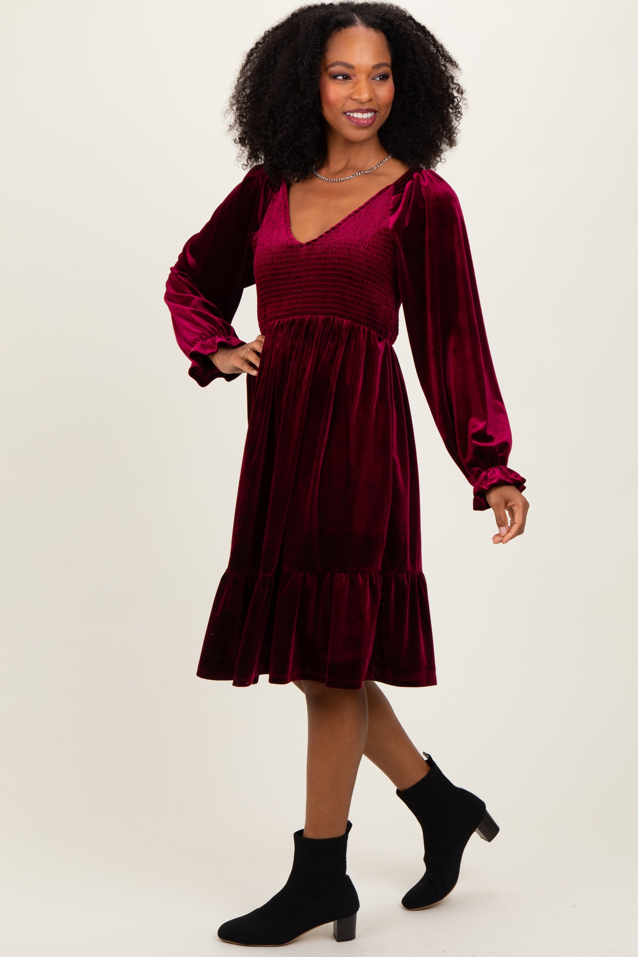 woman wearing a burgundy velvet dress with long sleeves, smocked bodice, and a ruffled hem, posing with a smile