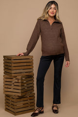 woman wearing a dark brown zip-up maternity sweater and black maternity pants, posing with wooden crates