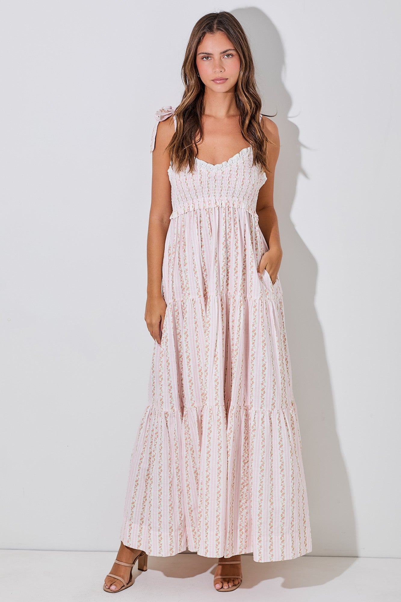 woman wearing a pink floral maxi dress with smocked bodice and tiered skirt, standing with hands in pockets