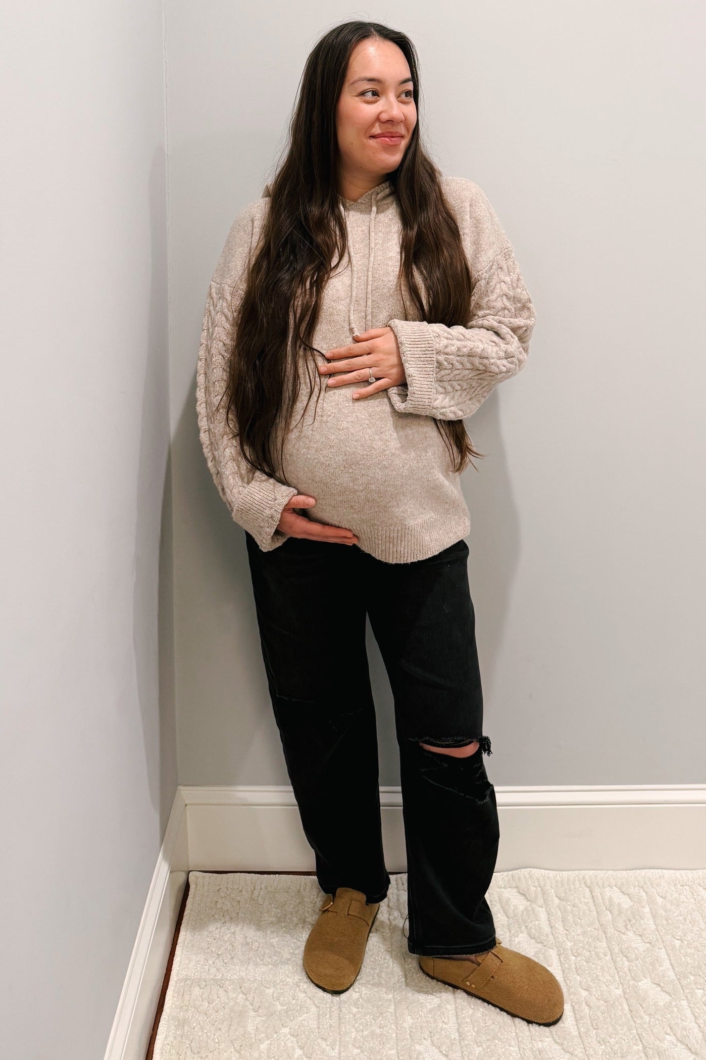 woman wearing a cream cable knit maternity hoodie and black ripped maternity jeans, holding her baby bump