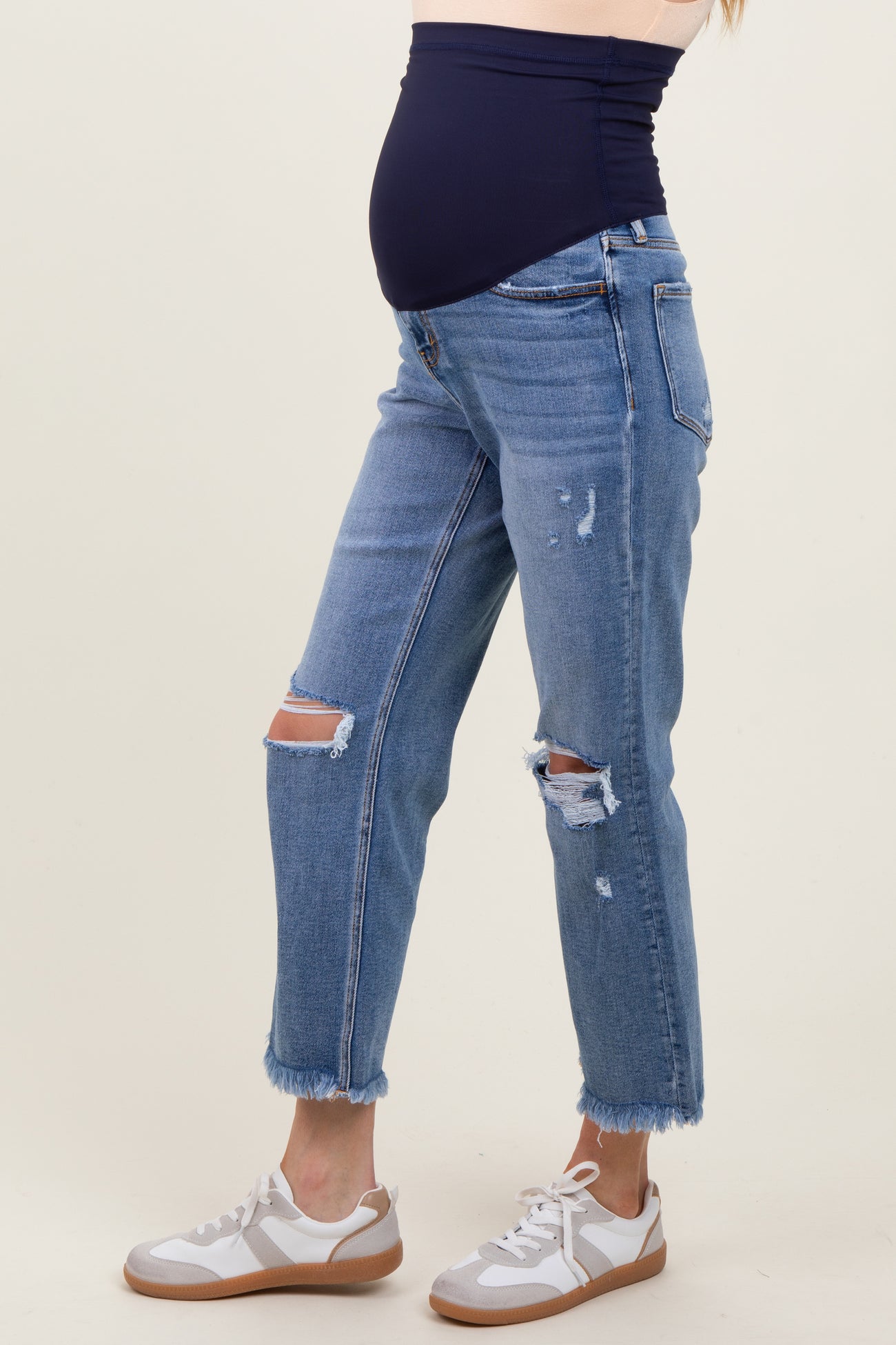 woman wearing distressed light blue maternity jeans with a navy belly panel and white sneakers, showcasing a baby bump