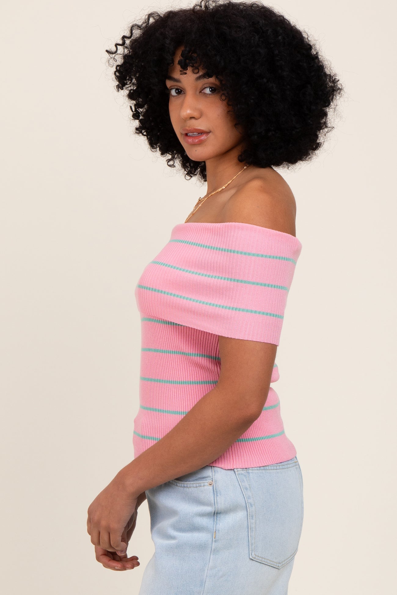woman wearing a pink off-the-shoulder ribbed top with teal stripes and light blue high-waisted jeans
