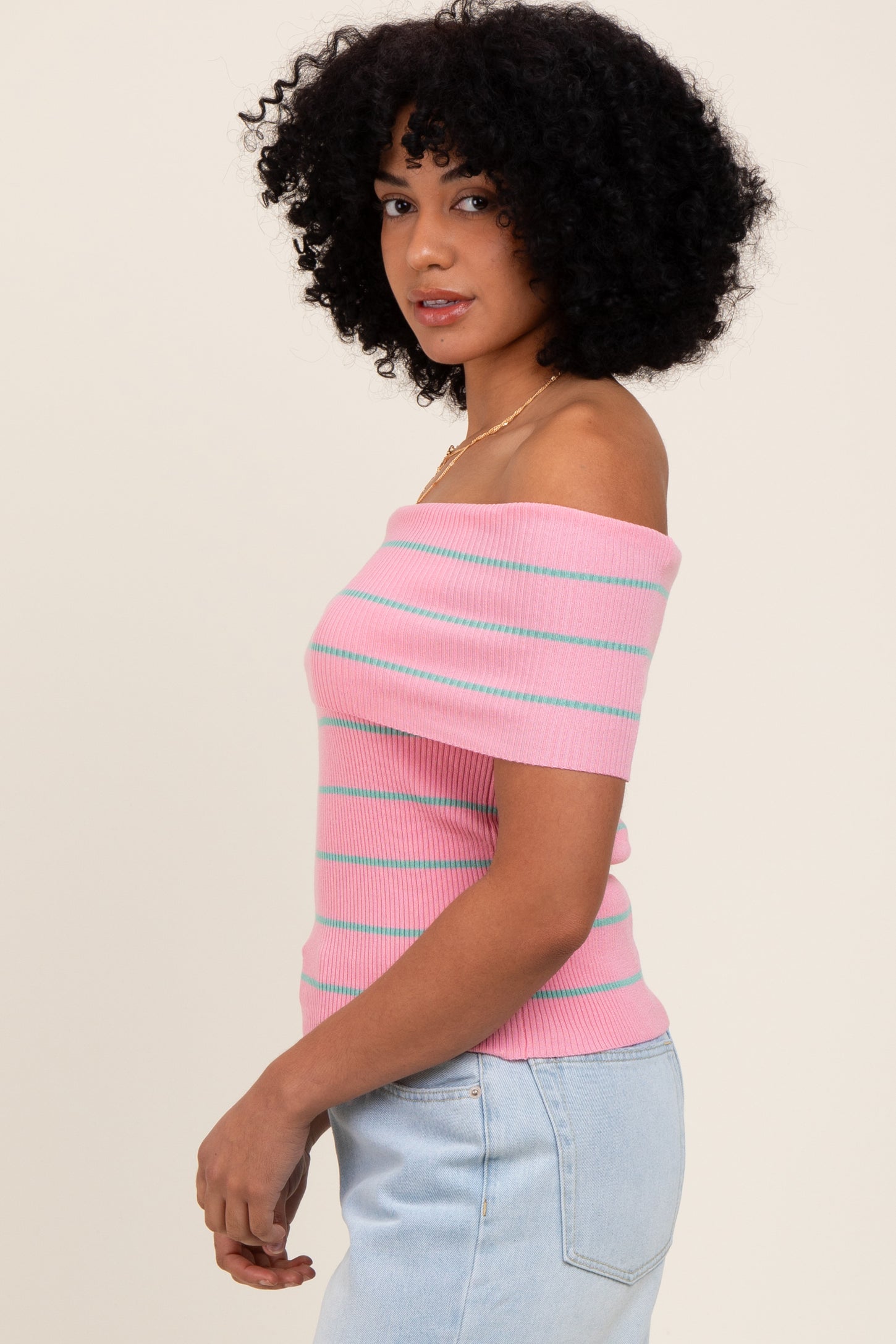 woman wearing a pink off-the-shoulder ribbed top with teal stripes and light blue high-waisted jeans