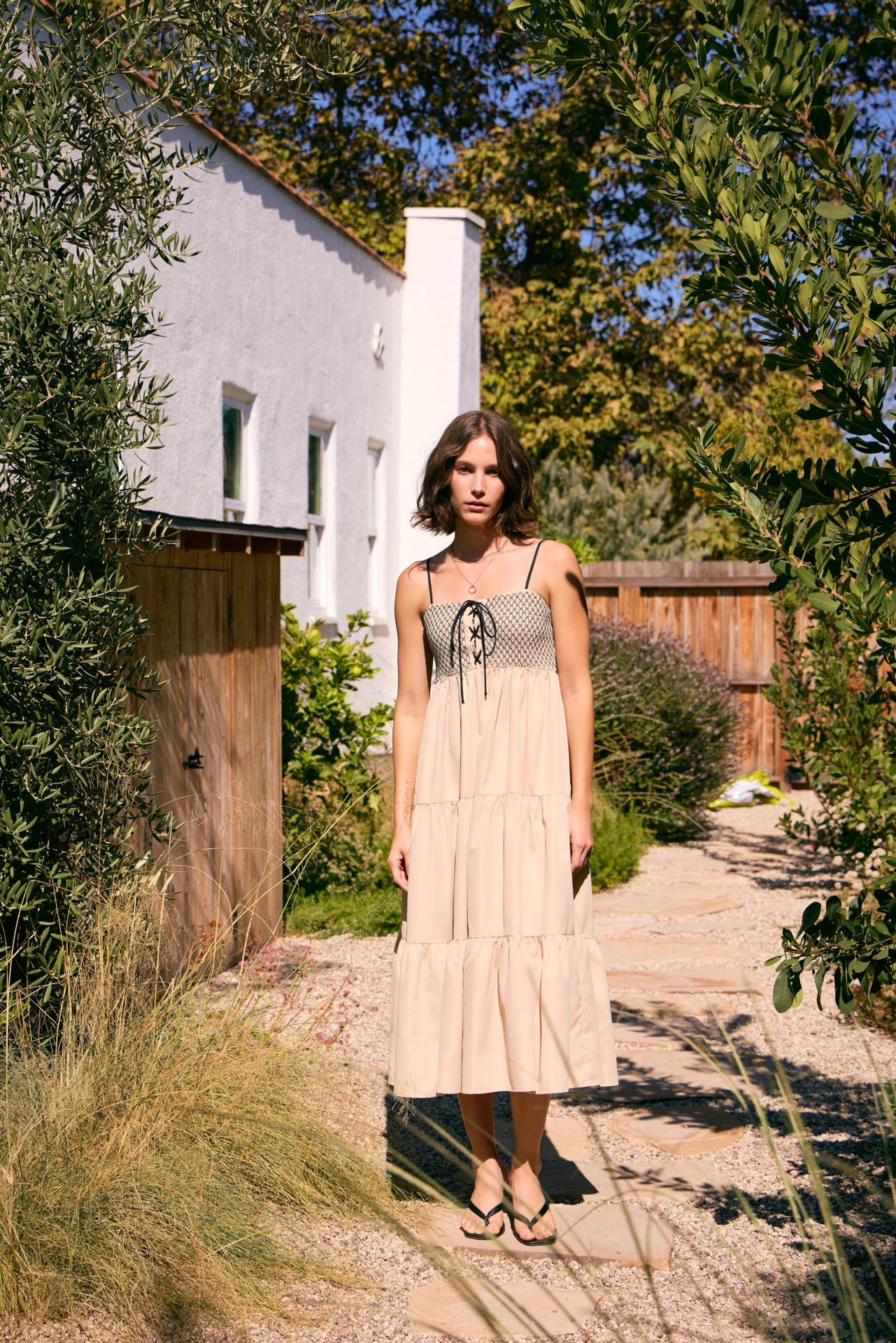 woman wearing a beige tiered maxi dress with a checkered bodice and adjustable tie detail, standing in a garden setting