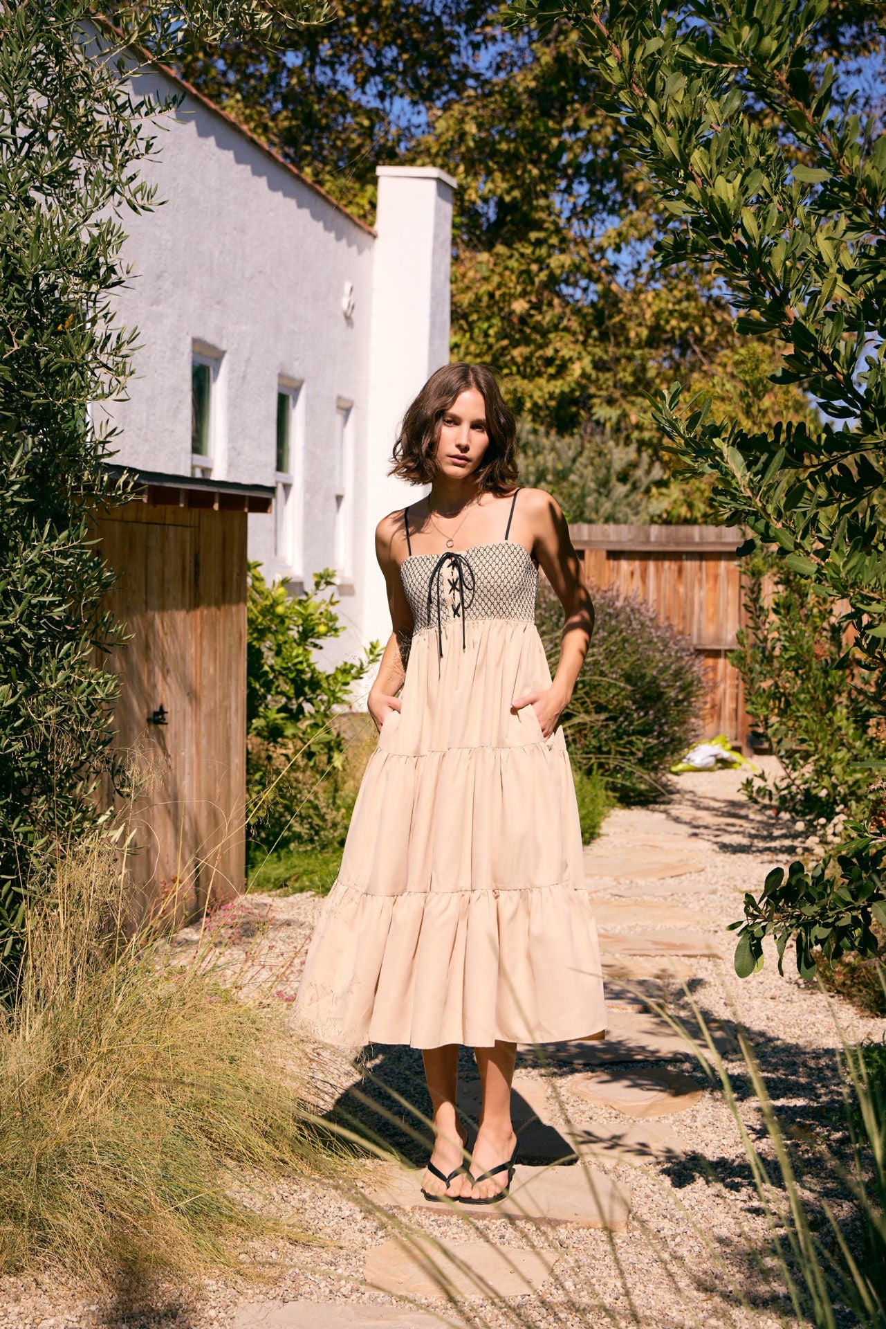 woman wearing a beige maxi dress with a smocked bodice and pockets, standing in a garden path