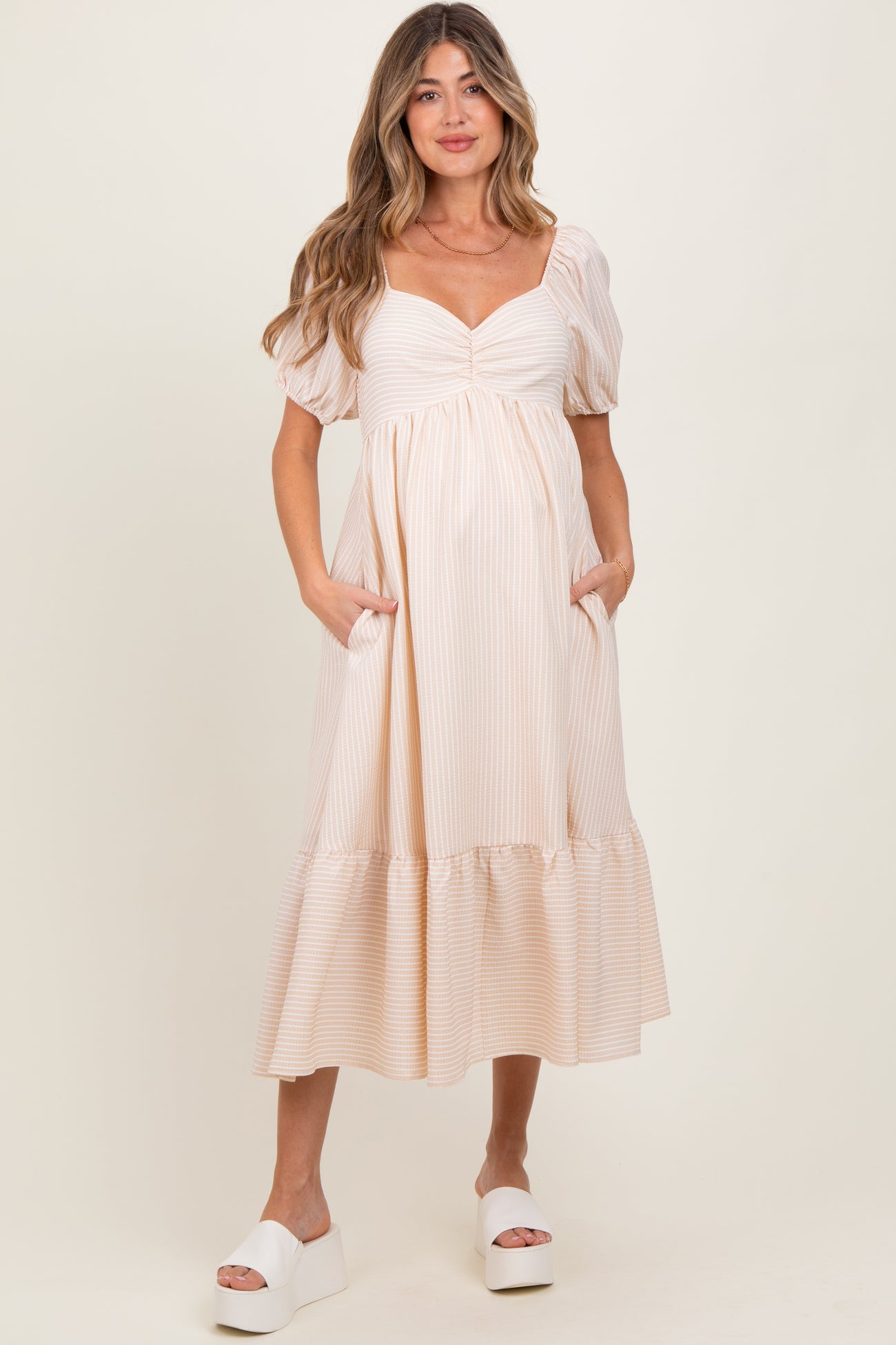 woman wearing a striped beige maternity dress with puff sleeves and pockets, standing and smiling