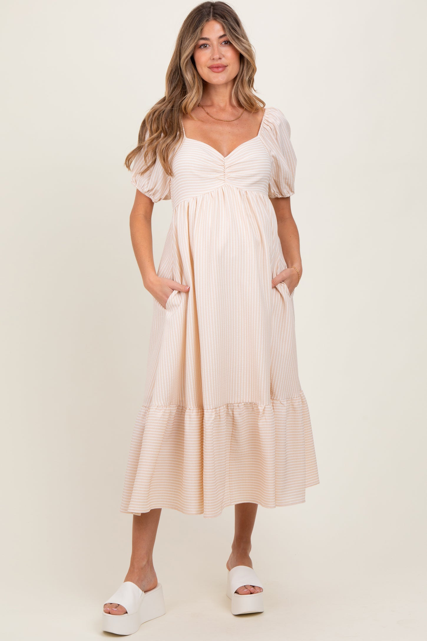 woman wearing a striped beige maternity dress with puff sleeves and pockets, standing and smiling