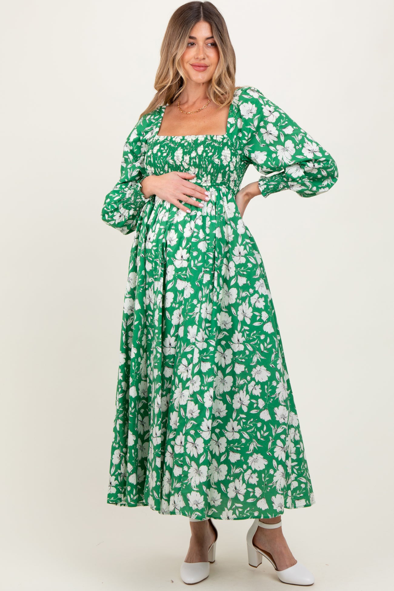 woman wearing a green floral maternity maxi dress with long sleeves and a smocked bodice, holding her baby bump