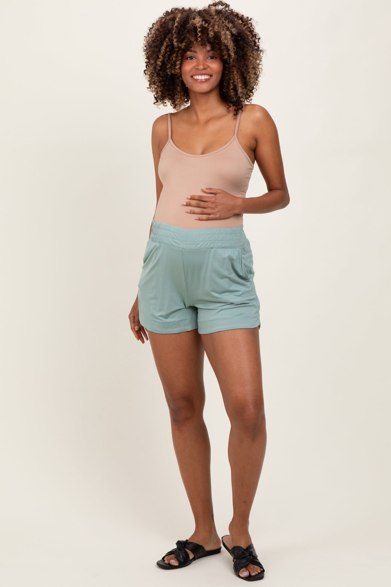 woman wearing a beige maternity tank top and teal maternity shorts, holding her baby bump