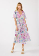 woman wearing a long, flowing floral maxi dress in pastel shades of purple, pink, and blue with short sleeves