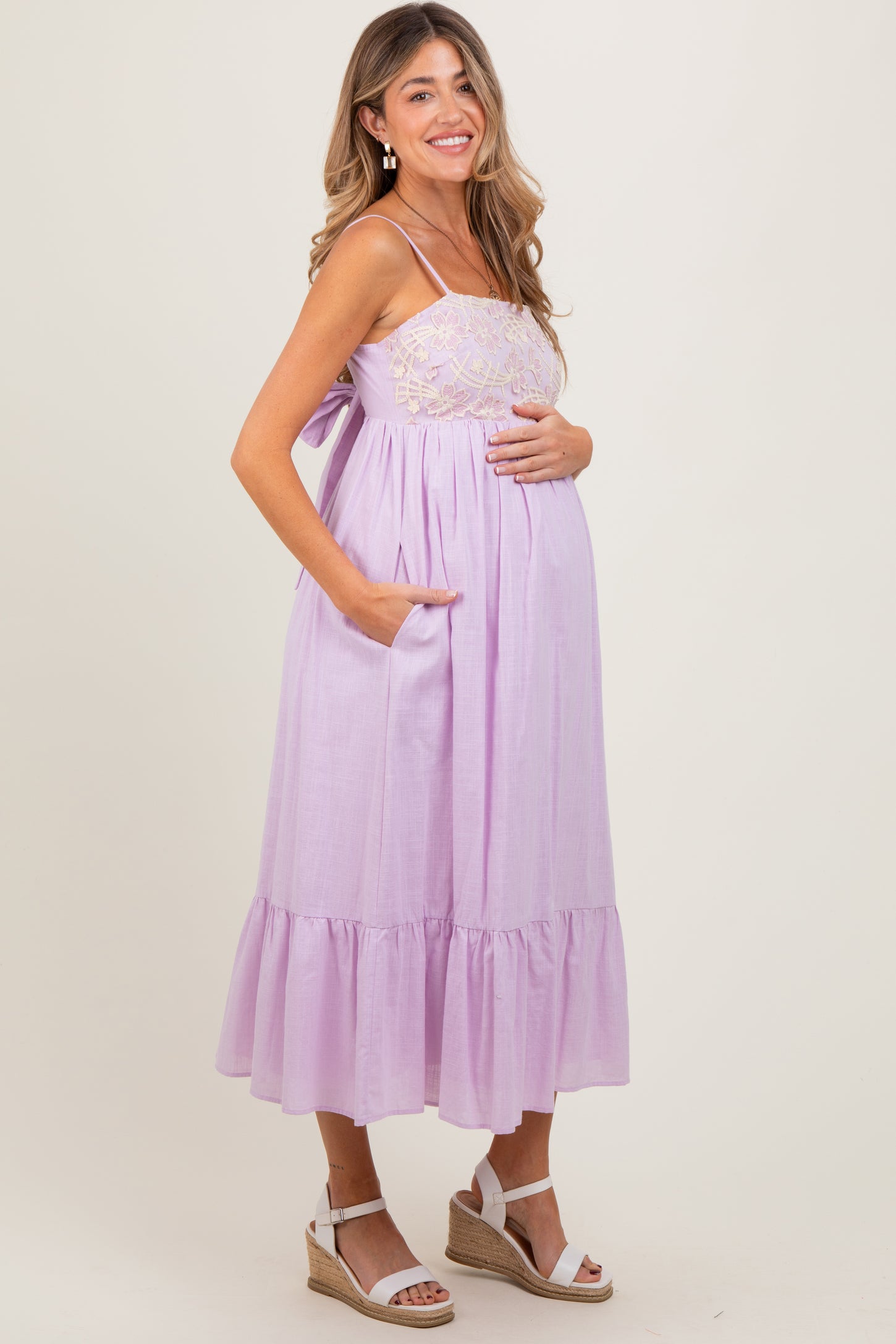 woman wearing a lavender maternity dress with embroidered details and pockets, holding her baby bump