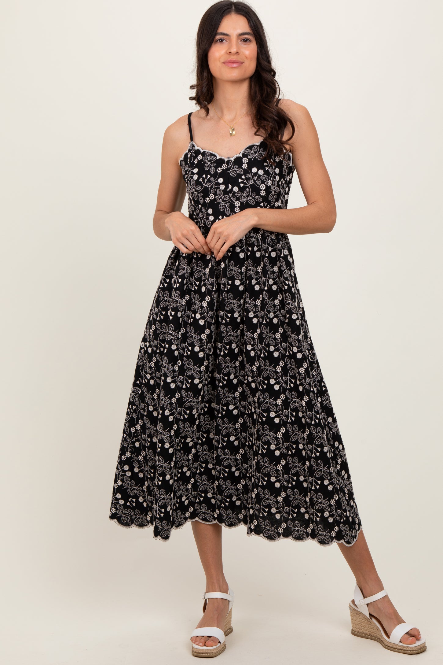 woman wearing a black floral midi dress with spaghetti straps and scalloped hem, standing with one hand on her hip
