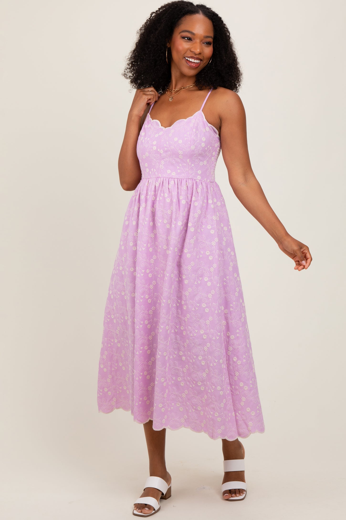 woman wearing a lilac midi dress with a scalloped neckline and floral details, styled with white sandals