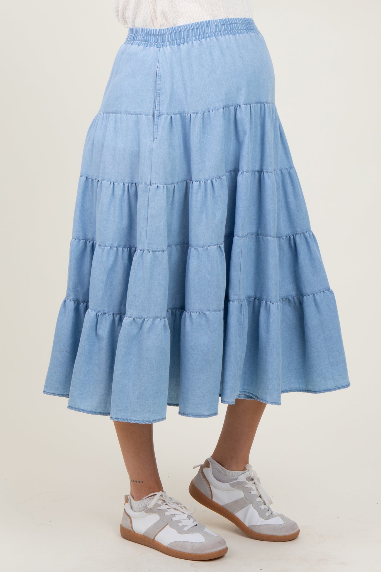 woman wearing a light blue tiered denim maternity skirt with an elastic waist and white sneakers