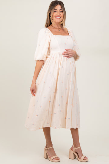 woman wearing a light pink maternity dress with puff sleeves and floral embroidery, holding her baby bump