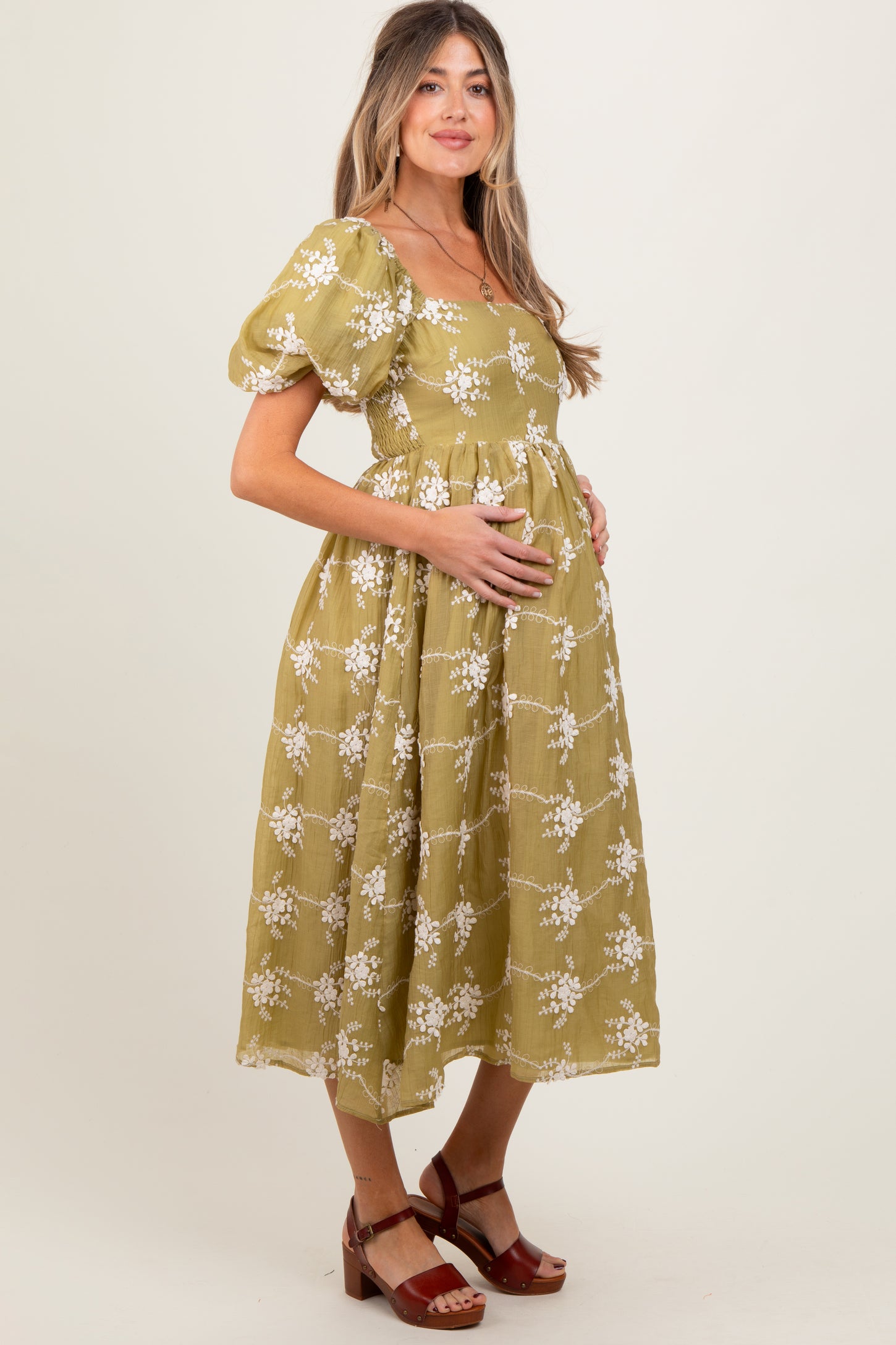 woman wearing a knee-length olive green maternity dress with puff sleeves and white floral patterns, holding her baby bump